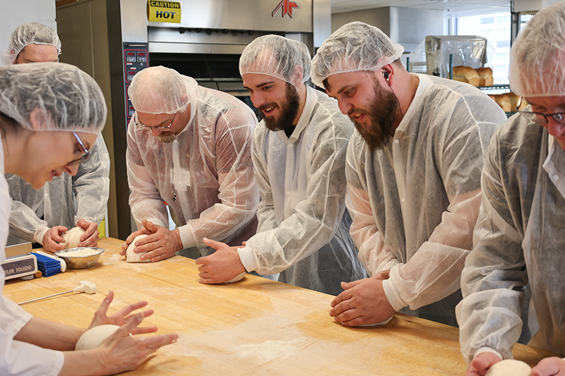 A Cereals Canada baking expert teaches participants to shape bread dough on a wooden table. A Cereals Canada baking expert teaches participants to shape bread dough on a wooden table.